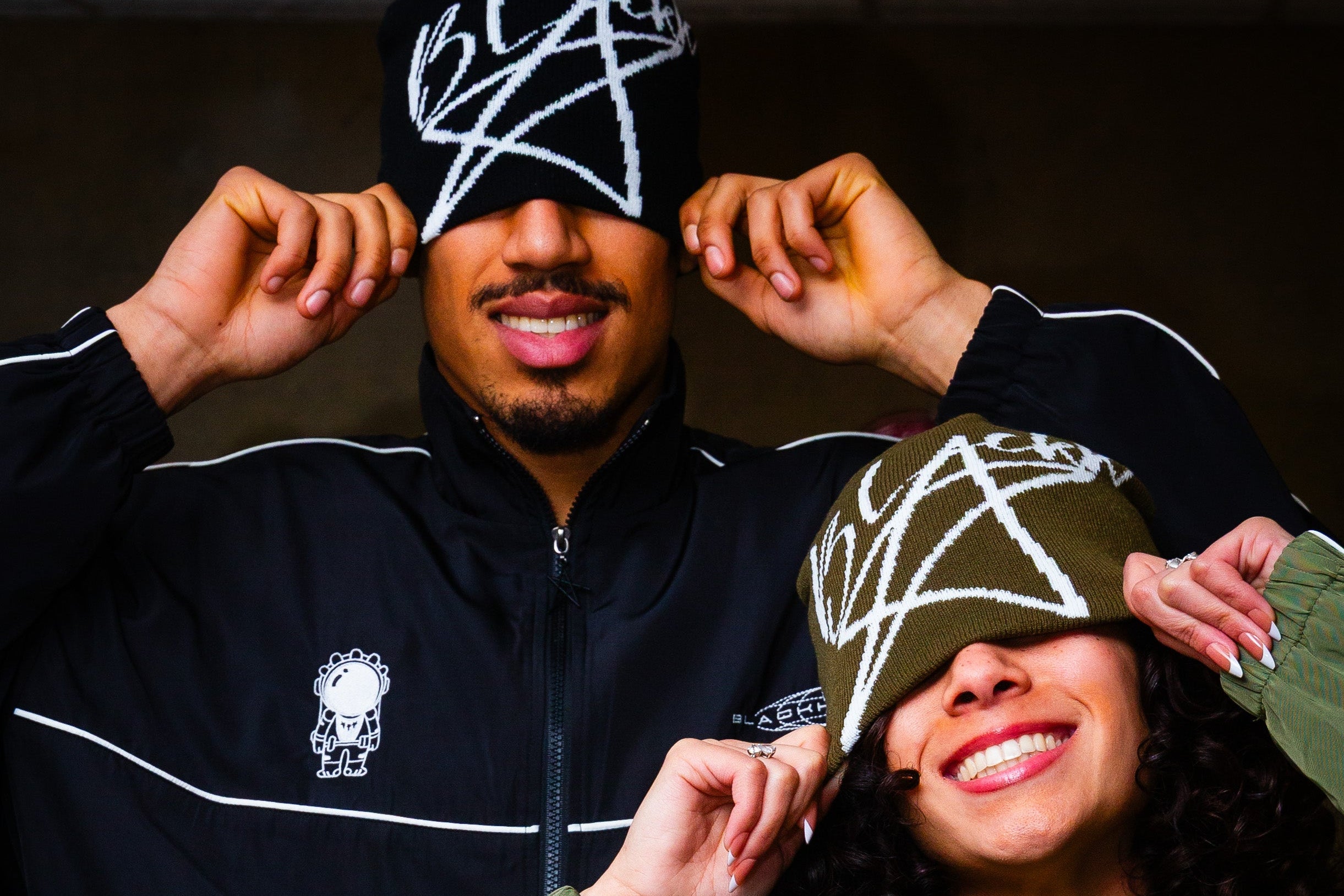 Two people wearing matching jackets with a logo, posing against a dark background.